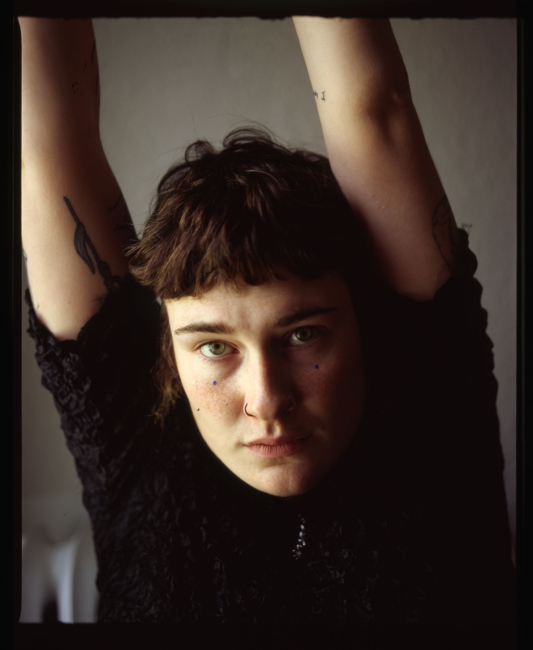 A photo of Reilly, their arms are stretched out above their head. They are gazing into the camera with a gentle, yet serious gaze. 